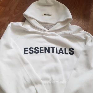 Vit Essentials Fear of God hoodie S - Säljer en clean vit hoodie från Essentials Fear of God i storlek S. Hoodien har svart tryck med ESSENTIALS på bröstet och är tillverkad i mjuk bomullsmix. Klassisk huva och ribbade muddar, perfekt för en chill och stilren look.