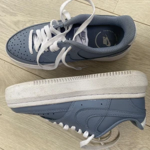 Nike Air Force 1 blå sneakers - Snygga Nike Air Force 1 sneakers i en ljusblå färg med en hög vit sula och vita skosnören. Skorna har klassisk rund tå, perforerad tåbox och är tillverkade i skinn. Perfekt för dig som gillar stilrena och tidlösa sneakers med en modern touch. Aldrig använda och ordinarie pris 1000kr 