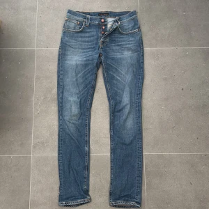 Blå jeans från Nudie Jeans - Säljer ett par klassiska blå jeans från Nudie Jeans med orange kontrastsömmar och snygg broderad detalj på bakfickorna. Jeansen har normal passform, raka ben och knappgylf. Perfekta för dig som gillar stilrena och tidlösa jeans. W32 L3. Modell: GrimTim