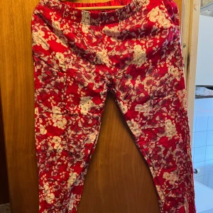 Röda blommiga byxor från Gina Tricot - Säljer ett par röda byxor från Gina Tricot med blommigt mönster i vitt och rosa. Byxorna har resår i midjan och är tillverkade i ett mjukt, lätt tyg. Perfekta för dig som vill ha något färgglatt och bekvämt.
