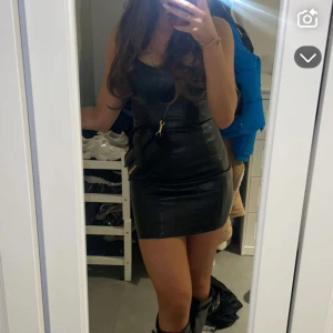 Svart tight kort klänning i skinnimitation - Säljer en svart, tight och kort klänning i skinnimitation med ärmlös design och rund halsringning. Klänningen har en snygg, figurnära passform som ger en cool och edgy look. Perfekt för dig som vill sticka ut och känna dig extra snygg.