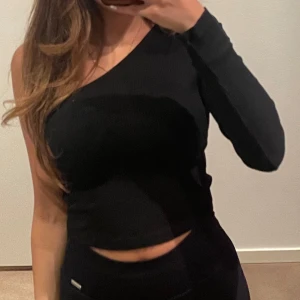 Svart oneshoulder topp croppad smalt ribbad - Snygg svart oneshoulder topp med lång ärm på ena sidan och croppad passform. Ribbad struktur som ger en cool vibe och passar perfekt till höga byxor. Enkel men ändå unik design som sticker ut.