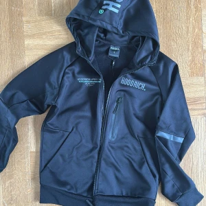 Svart hoodie från Hoodrich, S - Svart luvtröja från Hoodrich med huva och tryck på både bröstet och ryggen. Materialet är ganska glansigt. Jackan har dragkedja framtill, en bröstficka med dragkedja och ribbade muddar. Snygg streetstil med sportiga detaljer och logga på huvan. Köpt i London.
