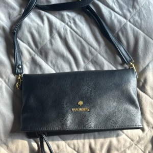 Svart axelväska från Mulberry i skinn - 4 olika fack går att vika ut eller vika in. Använd men fortfarande så fin och funktionell:) Säljer en stilren svart axelväska från Mulberry i äkta skinn med guldiga detaljer och logga framtill. Väskan har justerbar axelrem, dragkedja och innerfack med Mulberry-tryck. Perfekt storlek för det viktigaste och snygg till alla outfits.