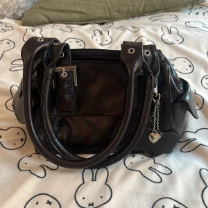 Mörkbrun handväska med hjärtberlock - Fin mörkbrun handväska i skinnliknande material. Väskan har tre stora fack och en hjärtberlock. Köpt på vinted 🩷