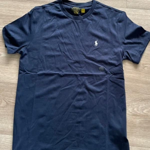 Mörkblå t-shirt Polo Ralph Lauren - Snygg mörkblå t-shirt från Polo Ralph Lauren i bomull med klassisk vit broderad logga på bröstet. Modellen är slim fit med rund hals och korta ärmar. Helt oanvänd endast testad. Har legat och skräpat i mitt rum det senaste året men skicket är fortfarande det samma som nytt! Pris kan även diskuteras vid snabb affär.