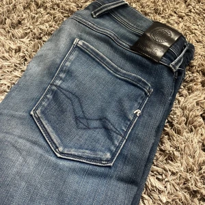 Replay Anbass blå jeans - Snygga Replay Anbass jeans i klassisk blå tvätt med slitningar och tydliga sömmar på bakfickan. Jeansen har normal passform, fem fickor och logga på bakre läderpatch. Perfekta för dig som gillar en stilren och modern look.