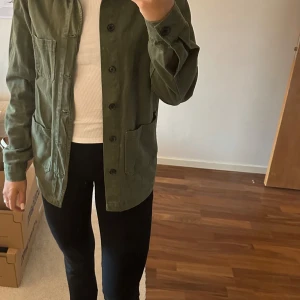 Grön overshirt från Roots - Säljer en grön overshirt från Roots i workwear-stil. Jackan har knäppning framtill, stora fickor och är långärmad. Perfekt att slänga över en t-shirt för en avslappnad look.