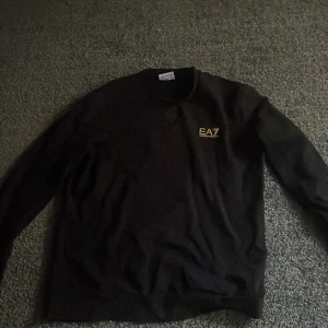 Svart sweatshirt från EA7 - Svart sweatshirt från EA7 med guldig logga på bröstet och gul detalj i nacken. Klassisk rund halsringning och långa ärmar. Perfekt för en stilren och sportig look.