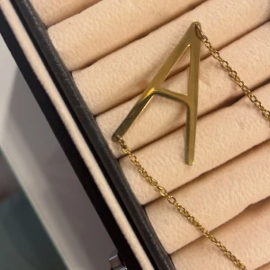 Guldfärgat halsband med bokstaven A - Stilrent halsband i guldfärg med en stor, minimalistisk bokstav A som hänge. Kedjan är tunn och klassisk, perfekt för dig som vill ha en personlig touch på din look. Passar lika bra till enkel t-shirt som till en snygg skjorta.