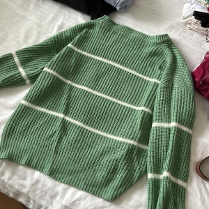 Grön randig stickad tröja - Snygg stickad tröja i grönt med vita horisontella ränder. Tröjan har en grovstickad struktur, långa ärmar och ribbade muddar vid ärmslut och nederkant. Perfekt för dig som gillar en chill och trendig look.