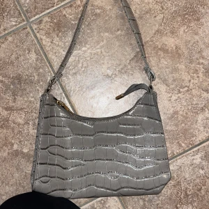 Grå axelväska i krokopräglat skinn - Snygg grå axelväska med croco-mönster och glansig finish. Väskan har en dragkedja upptill och ett stilrent, modernt utseende. Perfekt storlek för det viktigaste och enkel att slänga över axeln.