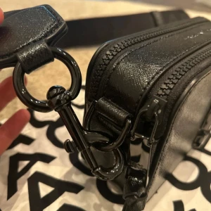 Svart axelväska Marc Jacobs - Snygg svart axelväska från Marc Jacobs i strukturerat skinn. Väskan har två dragkedjor, bred justerbar axelrem och metallogo framtill. Stilren och boxig form, perfekt för att bära det viktigaste. Diskreta svarta metalldetaljer och innerfack med logotyp.