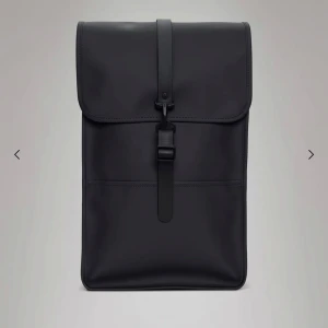 Svart ryggsäck från Rains - Snygg svart ryggsäck från Rains i stilren, fyrkantig design. Väskan har mattsvart finish, justerbara remmar och ett lock med spänne framtill. Baksidan har dragkedjeficka och remmar i syntetmaterial. Perfekt för dig som gillar clean och modern look.                                                          Standardstorleken, rymmer 13-tums laptop i laptopfickan.  30 cm x 48 cm x 12 cm