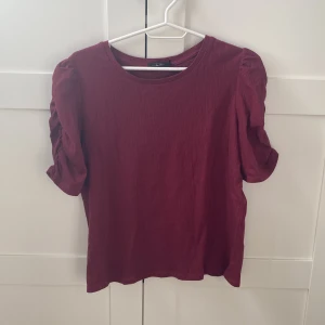 Vinröd topp med puffärmar - Säljer en vinröd topp med korta puffärmar och rund halsringning. Toppen har en enkel design med fina rynkade detaljer på ärmarna som ger en trendig look. Perfekt att styla till jeans eller kjol.