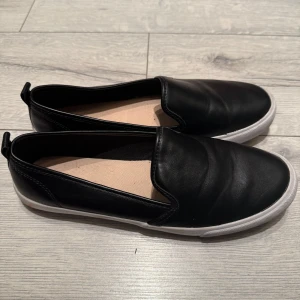 Svarta slip-on skor från H&M - 🌸Svarta slip-on skor från H&M med vit platt sula. Skorna har en enkel och stilren design i syntetmaterial. Ytterst lite använda, mycket bra skick 🌸