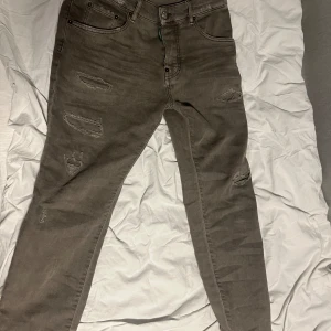 Grå slitna jeans från Dsquared2 - Säljer ett par grå jeans från Dsquared2 med slitningar på framsidan och klassisk femficksdesign. Jeansen har smal passform och detaljer som knappgylf och Dsquared2-lapp bak i midjan.