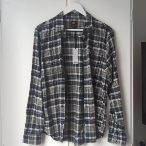 Rutig skjorta från Lee i bomull - Snygg rutig skjorta från Lee i regular fit, storlek M. Skjortan har gröna, svarta och vita rutor, klassisk krage och knäppning framtill. Tillverkad i mjuk bomull och har två bröstfickor med knappar. Perfekt för en avslappnad och cool stil.