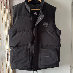 Svart dunväst från Canada Goose - Svart dunväst från Canada Goose med hög krage, två stora fickor framtill och broderad logotyp på bröstet. Västen har dragkedja och tryckknappar samt en klassisk, quiltad design. Perfekt för lager-på-lager under kyliga dagar.