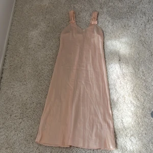 Ljusrosa långklänning med rosetter - Superfin långklänning i ljusrosa med breda axelband och söta rosetter på axlarna. Klänningen har en lätt glansig yta och v-ringning framtill. Perfekt för sommarens alla tillfällen när du vill känna dig extra söt.