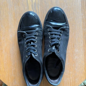 Lanvins  - Snygga svarta sneakers från Lanvin med ovandel i mocka och lackad tå. Klassisk låg modell med snörning och rund tå. Sulan är i svart gummi och skon har diskreta metalldetaljer vid snörningen. Perfekta för en clean och stilren look. Dessa är i toppen skick och mockan är som ny. Det enda är själva tån som tagit lite skada men inget som syns vid användning. Storlek 42 