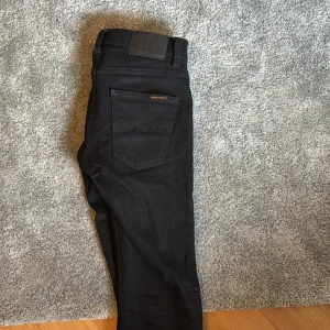 Svarta jeans från Nudie Jeans - Säljer ett par svarta jeans från Nudie Jeans med klassisk femficksdesign och smal passform. Jeansen har svarta sömmar, svarta nitar och läderpatch bak i midjan. Perfekta för dig som gillar stilrena och tidlösa byxor. Använda max 5 gånger.