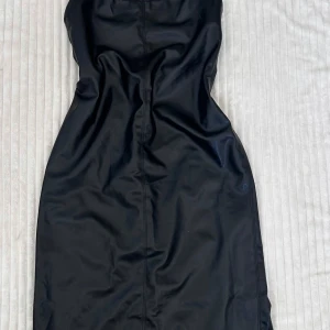 Svart långklänning i skinnimitation - Säljer en svart långklänning i skinnimitation med halterneck och öppen rygg. Klänningen har en tight passform och en stilren look som passar perfekt när du vill sticka ut. Snygg och trendig modell med slät yta och snygga sömdetaljer.