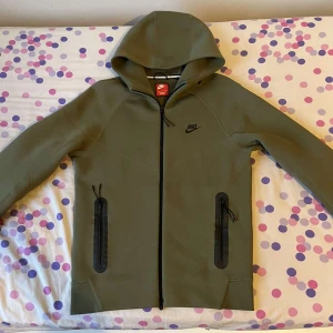 Olivgröna mjukisbyxor från Nike - Snygga olivgröna mjukisbyxor från Nike med svarta dragkedjor på fickorna och resår i midjan. Byxorna har en modern, avsmalnande passform och är perfekta för en avslappnad stil. Nike-logga på låret.