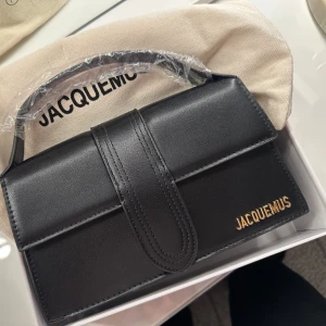 Väska Jacquemus - Säljer en stilren svart handväska från Jacquemus med guldfärgad logga. Väskan har ett elegant, fyrkantigt format. Går att använda som handväska och axelremsväska. Den är helt oanvänd. Kommer med dustbag