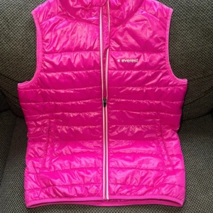 Rosa dunväst från Everest - Snygg rosa dunväst från Everest med quiltad design och hel dragkedja framtill. Västen har hög krage och två fickor på sidorna. Perfekt för lager-på-lager och ger en poppig färg till din outfit.                                                                    Jackan passar även XS/S