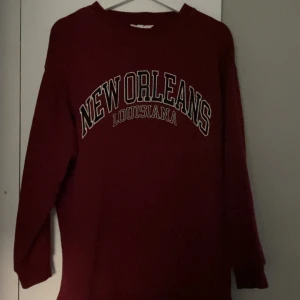 Vinröd sweatshirt New Orleans - Vinröd sweatshirt med trycket 'New Orleans Louisiana' i vitt och svart på bröstet. Klassisk rund hals och långa ärmar, perfekt för en chill och avslappnad stil. Passar dig som gillar collegevibbar och streetwear.