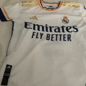 Real Madrid Bellingham matchtröja - Säljer en officiell Real Madrid fotbollströja från Adidas med Bellingham och nummer 5 på ryggen. Tröjan är vit med guldiga detaljer på axlarna, mörkblå tryck och inslag av orange vid krage och ärmslut. Materialet är lätt och andas, perfekt för fotboll eller supporterliv.