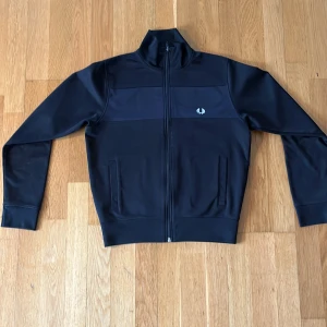Svart track jacket från Fred Perry - Tjena! Säljer en snygg svart track tjocktröja från Fred Perry med den klassiska broderade loggan på bröstet. Materialet är mjukt och skönt. Perfekt för en sportig och clean look.