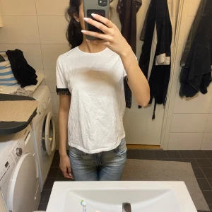 Vit t-shirt med svarta spetsdetaljer - Vit t-shirt med korta ärmar och snygga svarta spetsdetaljer längst ut på ärmarna. Modellen är rak och lite loose fit, perfekt till jeans eller kjol. T-shirten är i mjuk bomull och har en enkel, clean look med en twist tack vare spetsen.