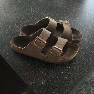 Birkenstocks - Säljer ett par klassiska bruna sandaler från Birkenstock med två justerbara remmar och metallspännen. Modellen har en anatomiskt formad fotbädd och platt sula för extra komfort. Perfekta för sommaren!