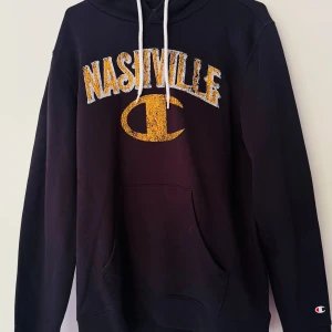 Svart hoodie från Champion med Nashville-tryck - Svart hoodie från Champion i storlek M med broderat gult och vitt Nashville-tryck på bröstet. Klassisk känguruficka framtill och vita dragsnören i huvan. Perfekt för dig som gillar streetwear och sportig stil.