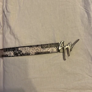 Svart och vit bälte från Giuseppe Zanotti med silverfärgat spänne - Säljer ett snyggt bälte från Giuseppe Zanotti i svart och vitt med paisleymönster och ett stort, silverfärgat signaturspänne. Bältet är gjort i skinn och har en exklusiv känsla, perfekt för att lyfta din outfit. Sälja väldigt billigt eftersom jag är sugen på ett annat bälte. Köpt runt 2000-2500kr.