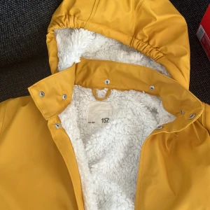 Gul fodrad regnjacka med huva - Säljer en gul regnjacka med vit teddyfoder och huva. Jackan har tryckknappar framtill och två stora fickor. Perfekt för kyliga och regniga dagar. Fodret är mjukt och värmande.