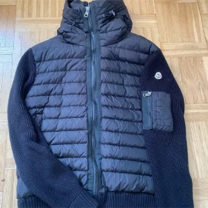 Svart pufferjacka från Moncler - Säljer en svart pufferjacka från Moncler med stickade ärmar och huva. Jackan har dragkedja framtill, en ficka med dragkedja på ärmen och Moncler-logga. Perfekt för sommarkvällar och snygg till vardags.