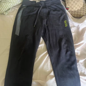 Svarta mjukisbyxor från Hugo Boss - Mörkblå mjukisbyxor från Hugo Boss med neon grön logotyp på benet och gråa detaljer längs sidorna. Byxorna har snörning i midjan och är perfekta för en avslappnad stil. Mjukt material och bekväm passform.