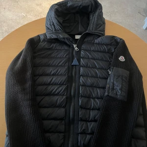 Svart Moncler pufferjacka med huva - Svart pufferjacka från Moncler med stickade ärmar och quiltad front. Jackan har huva, dragkedja framtill och en snygg Moncler-logga på ärmen. Materialmix av polyester och stickad ull ger både värme och stil. Perfekt för dig som vill ha en clean och modern look.