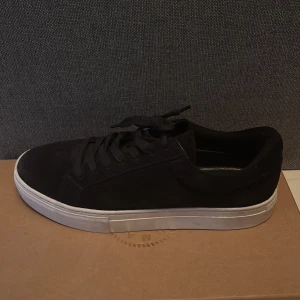 Svarta sneakers från Vagabond i mocka - Stilrena svarta sneakers från Vagabond med ovandel i mjuk mocka och klassisk vit platt sula. Skorna har rund tå, snörning och en clean look som passar till det mesta. Perfekta för dig som gillar minimalistisk stil och vill ha ett par bekväma vardagssneakers.