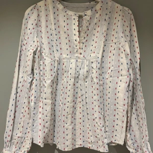 Skön långärmad blus - En fin och skön långärmad blus i bomull. Vit botten med ljusblå ränder och röd, blå detaljer. Knäppning framtill och broderad kant. Väldigt sparsamt använd, som ny. 