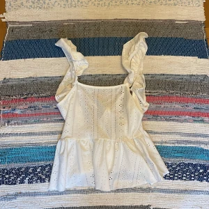 Vit volangtopp med broderade detaljer - Supersöt vit topp med breda volangaxelband och broderade hålmönster. Toppen har en luftig passform med volang nertill och knytning i ryggen. Perfekt för varma dagar när du vill känna dig extra gullig och somrig.