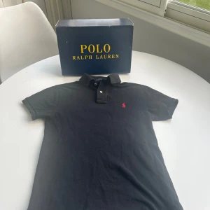 Svart pikétröja från Polo Ralph Lauren - Klassisk svart pikétröja från Polo Ralph Lauren i slim fit-modell. Tröjan har korta ärmar, krage med knappar och den ikoniska röda broderade loggan på bröstet. Perfekt för en stilren och sportig look.