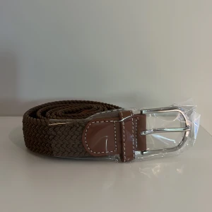Brunt flätat bälte  - Snyggt brunt flätat bälte med silverfärgat metallspänne och detaljer i brunt skinn. Perfekt accessoar för att lyfta din outfit. Klassisk design med vita sömmar på skinnpartierna.