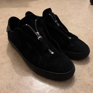 Svarta sneakers från Axel Arigato - Stilrena svarta sneakers från Axel Arigato i mocka med dragkedja framtill istället för klassisk snörning. Låg modell med rund tå och svarta sulor. Perfekta för dig som gillar minimalistisk och modern design. Skorna är sparsamt använda sedan jag köpt de. Ställ gärna frågor eller prisförslag.