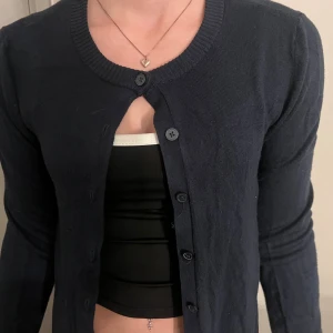 Mörkblå kofta med rund hals - En klassisk mörkblå kofta med rund halsringning och långa ärmar. Tröjan har knäppning framtill med svarta knappar och är tillverkad i ett mjukt, tunt material som passar perfekt till lager-på-lager-stil.