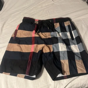 Rutig badshorts från Burberry - Säljer ett par snygga badshorts från Burberry med klassiskt rutigt mönster i svart, beige, vitt och rött. Shortsen har snörning i midjan och fickor på sidorna samt en bakficka. Perfekta för sommaren!