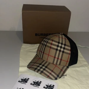 Rutig keps från Burberry - Snygg keps från Burberry med klassiskt rutigt mönster i beige, svart och rött. Framsidan är i bomullstyg och baksidan i svart mesh med justerbar rem. Perfekt accessoar för en trendig look.